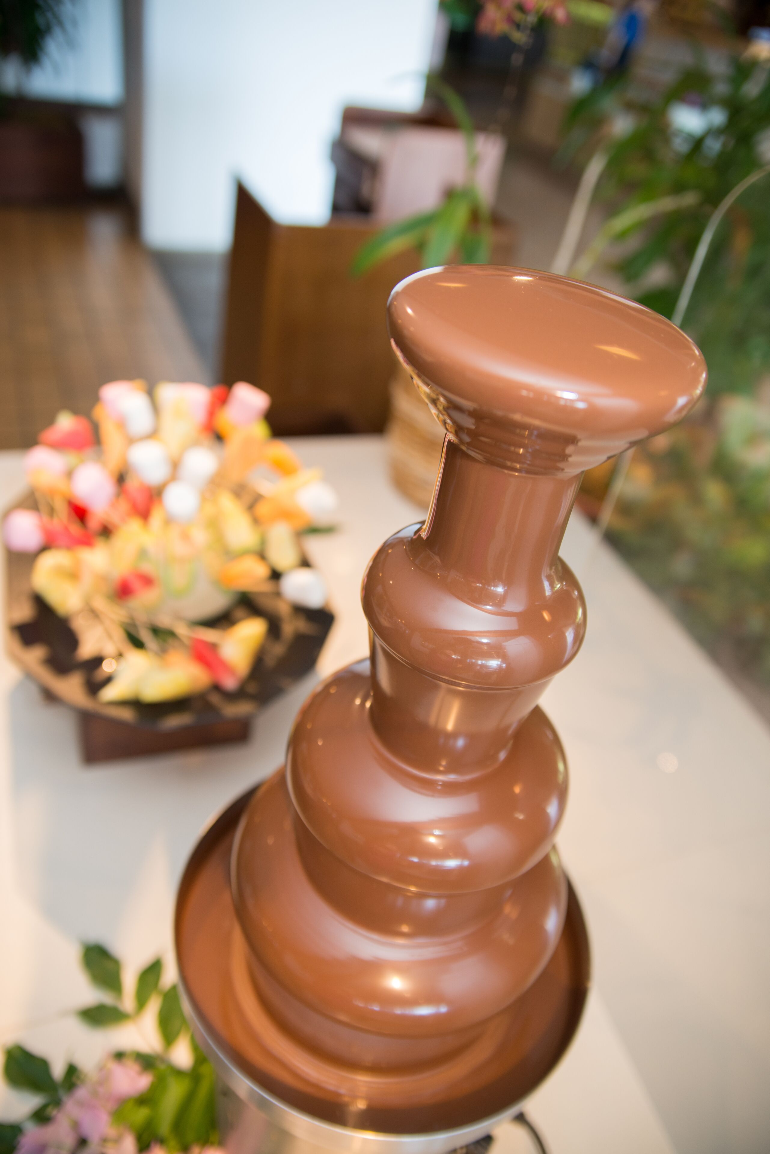 Eine dekorative Schokoladenbrunnen-Mitte auf einem Tisch, im Hintergrund eine Obstplatte.