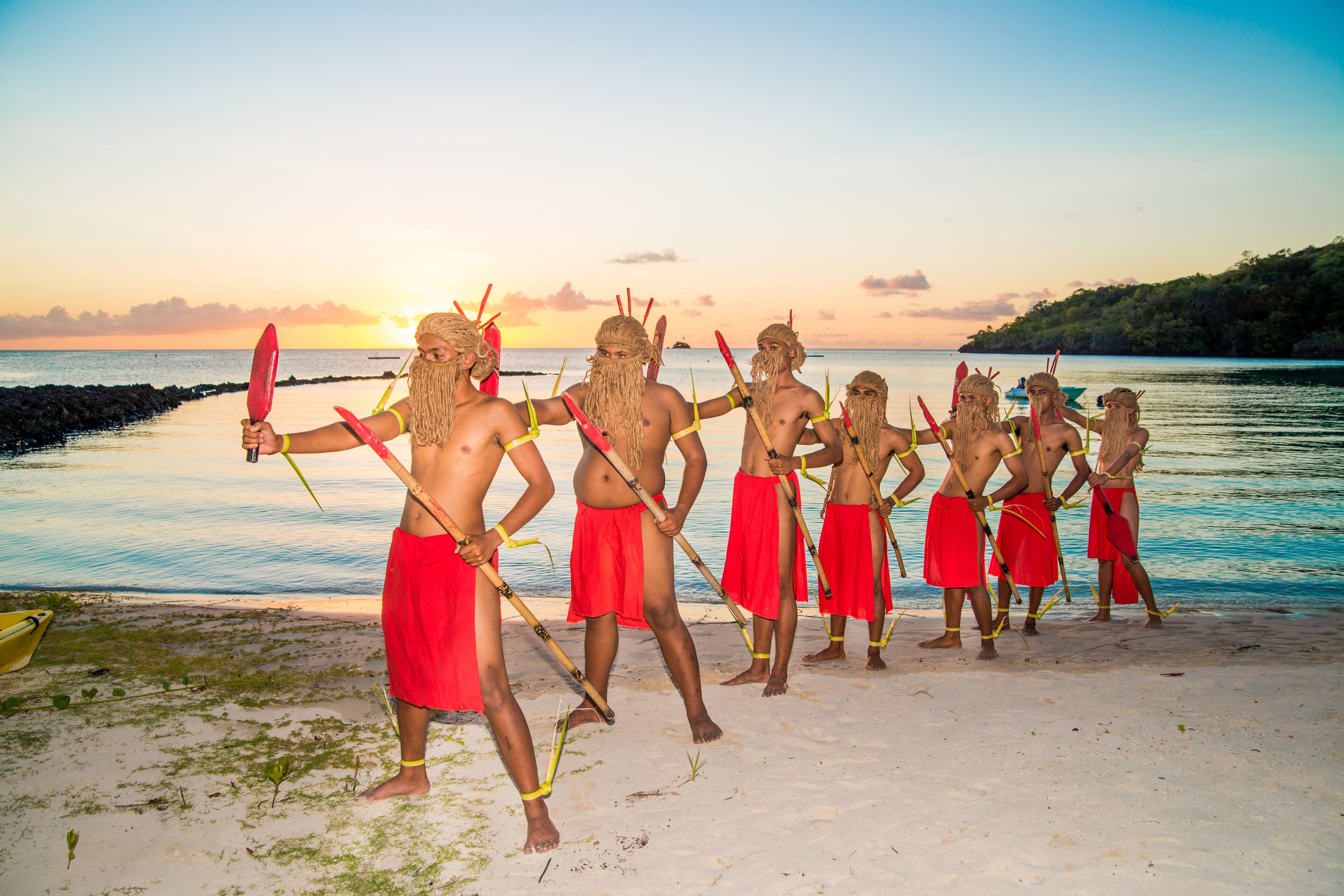 A group of people in traditional attire standing on a beach with a sunset in the background.