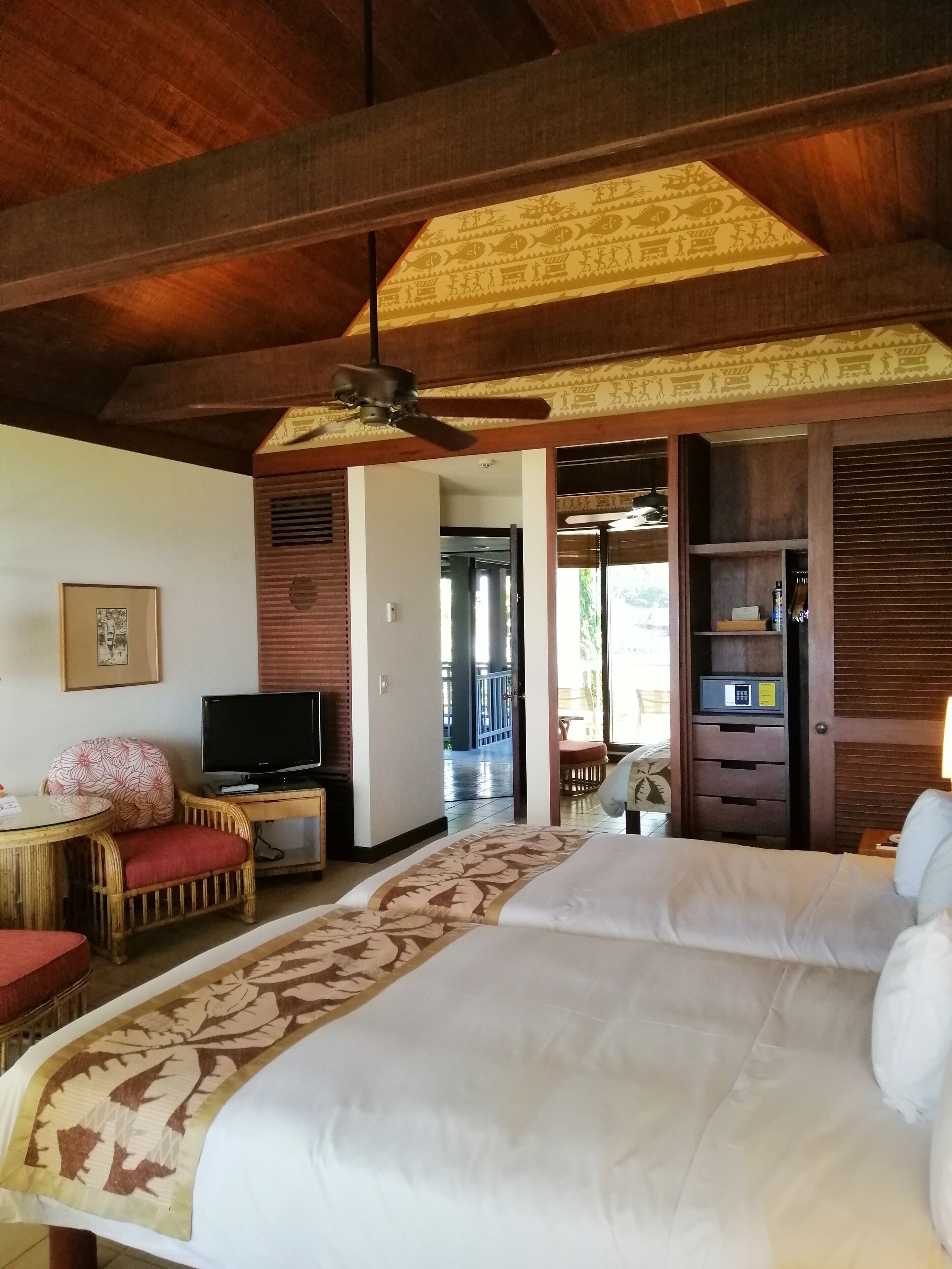 A hotel room with a bed, a television, a ceiling fan, a wooden wardrobe, and a small table with a chair.