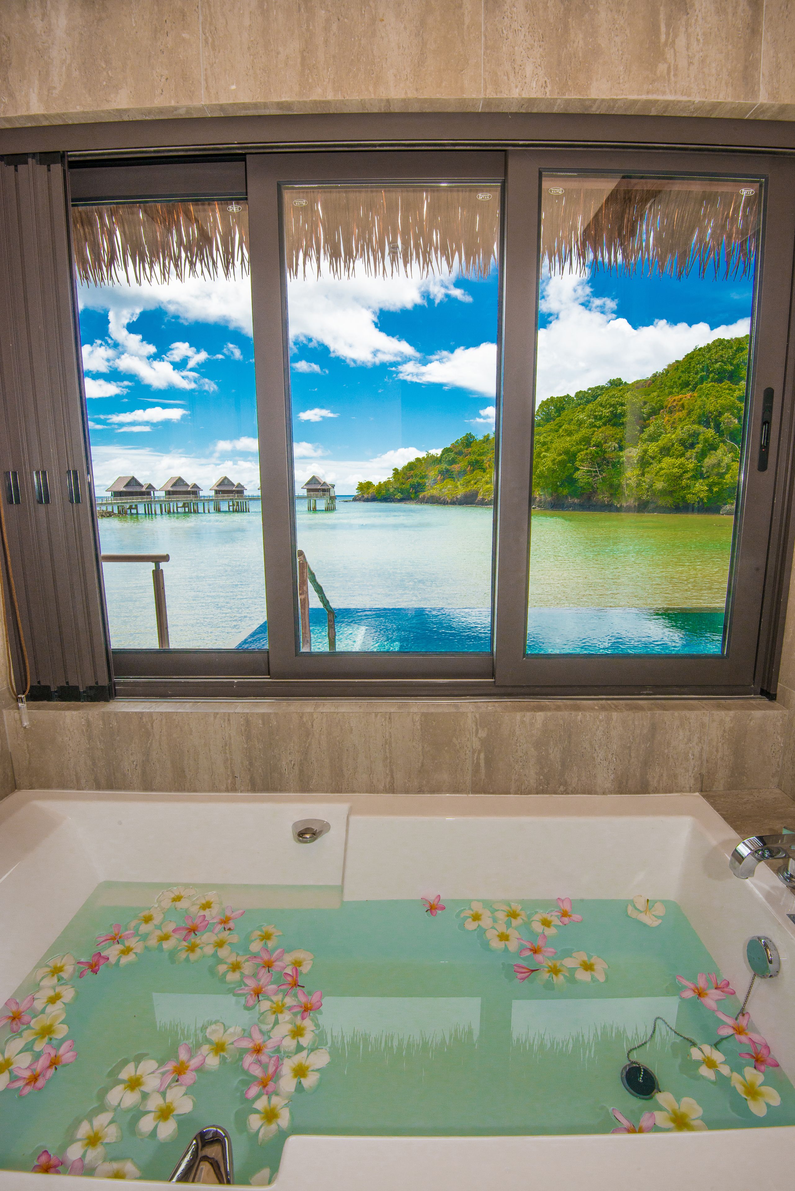A bathroom with a large window offering a view of a tropical beach with overwater bungalows.