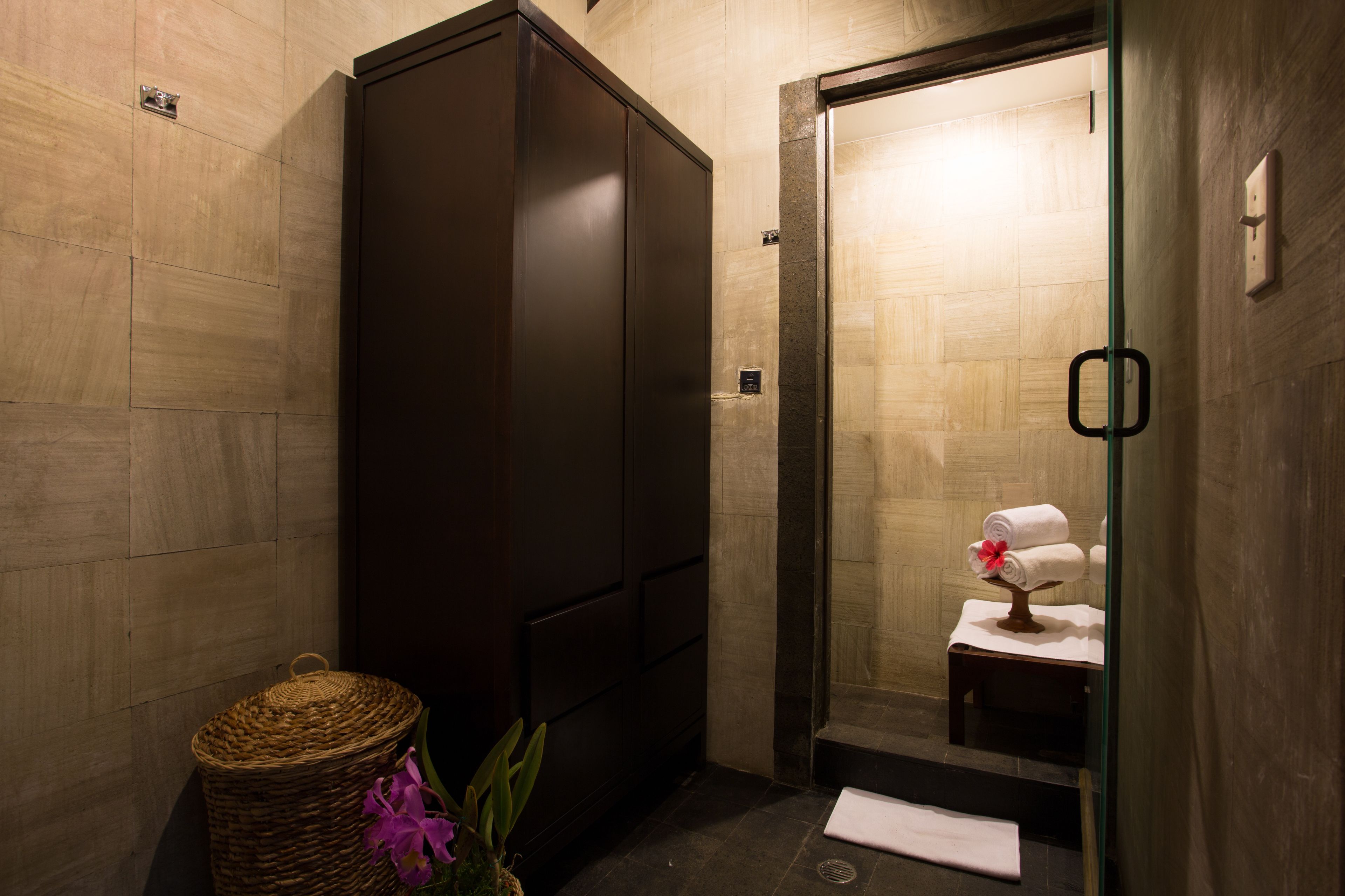 A bathroom with a dark wooden wardrobe, a wicker basket, and a small table with towels and a flower.