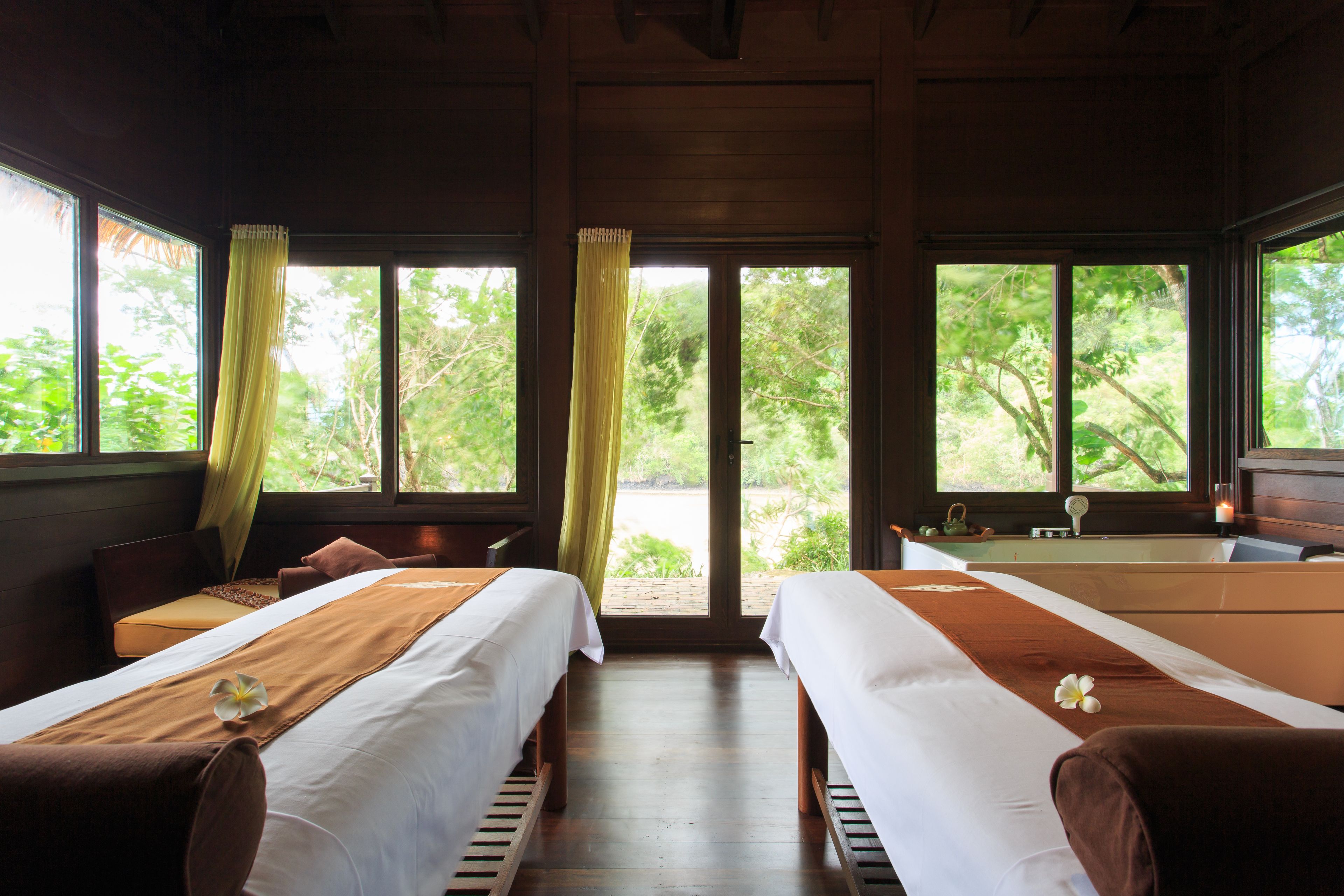 A room with two beds, a bench, and a view of greenery through large windows.