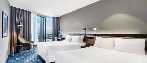 Een hotelkamer met twee bedden, een bureau, een stoel en een groot raam met uitzicht op de stad.