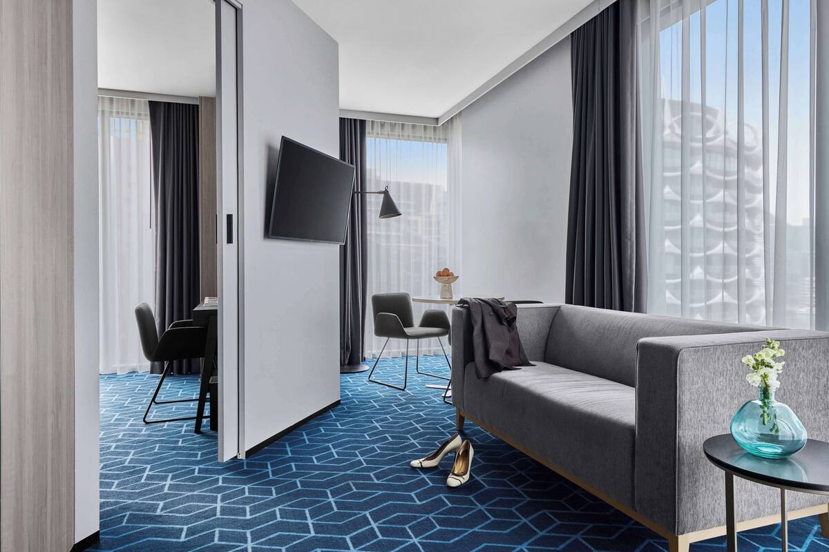 Ein modernes Hotelzimmer mit grauem Sofa, einem kleinen runden Tisch mit einer Vase, einem an der Wand befestigten Fernseher und einem blau gemusterten Teppich.