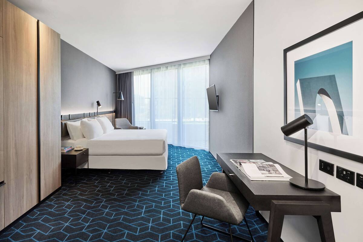 Ein modernes Hotelzimmer mit Bett, Schreibtisch, Stuhl, Fernseher und einem großen Fenster mit Vorhängen.