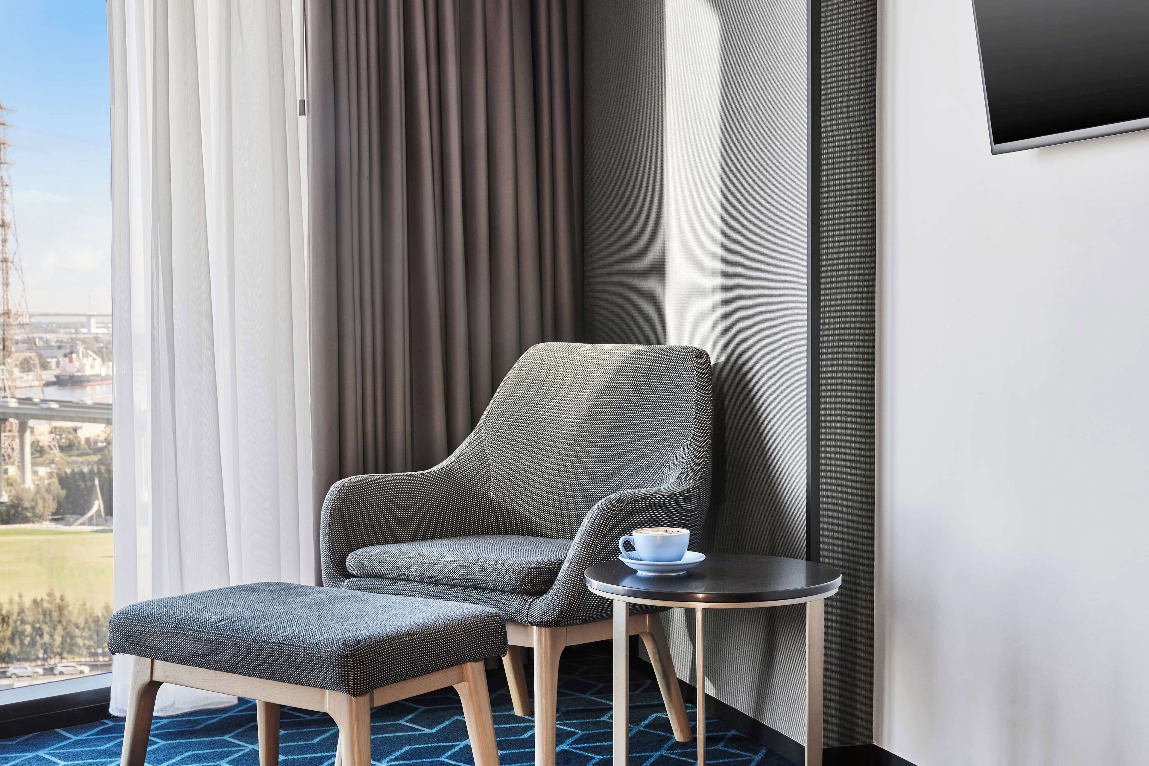 Una habitación de hotel moderna con un sillón tapizado gris, un reposapiés a juego y una pequeña mesa redonda con una taza.