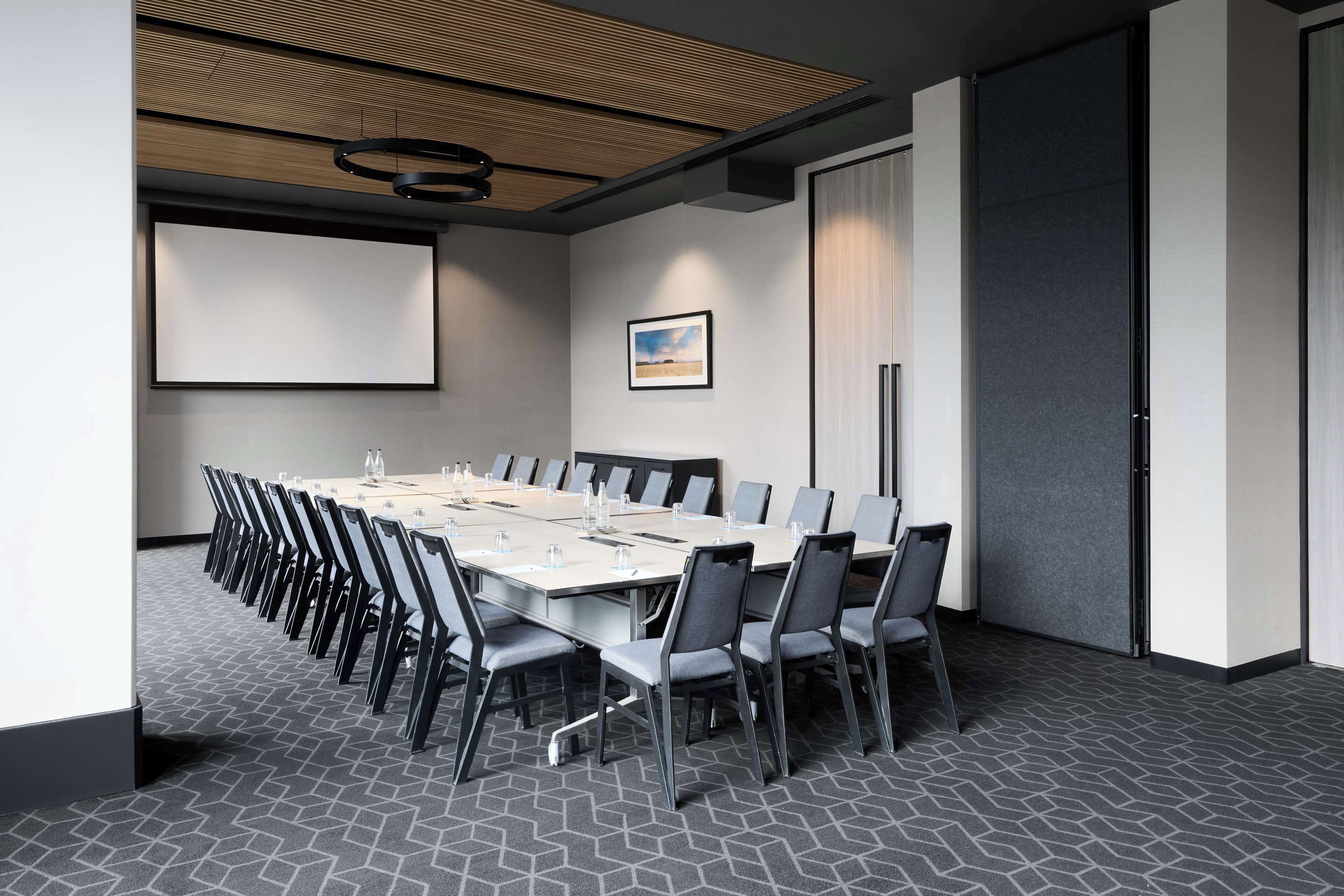 Ein Konferenzraum mit langem Tisch, Stühlen, einer Projektionsleinwand und einem gerahmten Bild an der Wand.
