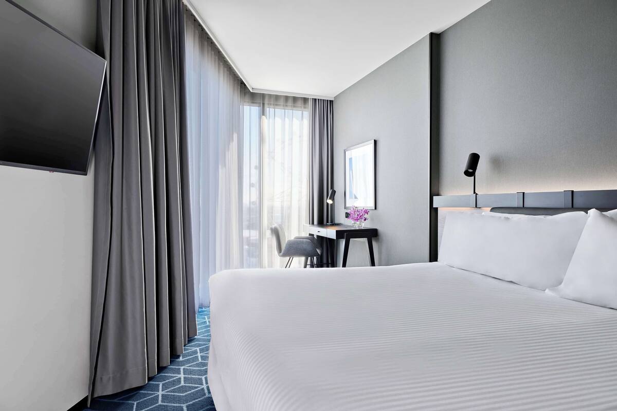 Ein Hotelzimmer mit einem großen Bett, einem Schreibtisch mit Stuhl, einem Flachbildfernseher und einem großen Fenster mit Vorhängen.
