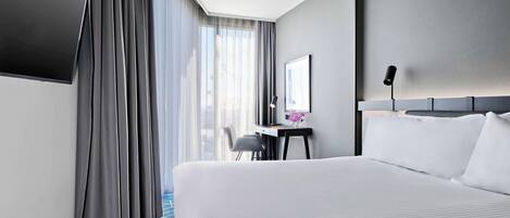 Una habitación de hotel con una cama grande, un escritorio con una silla, un televisor de pantalla plana y un ventanal con cortinas.