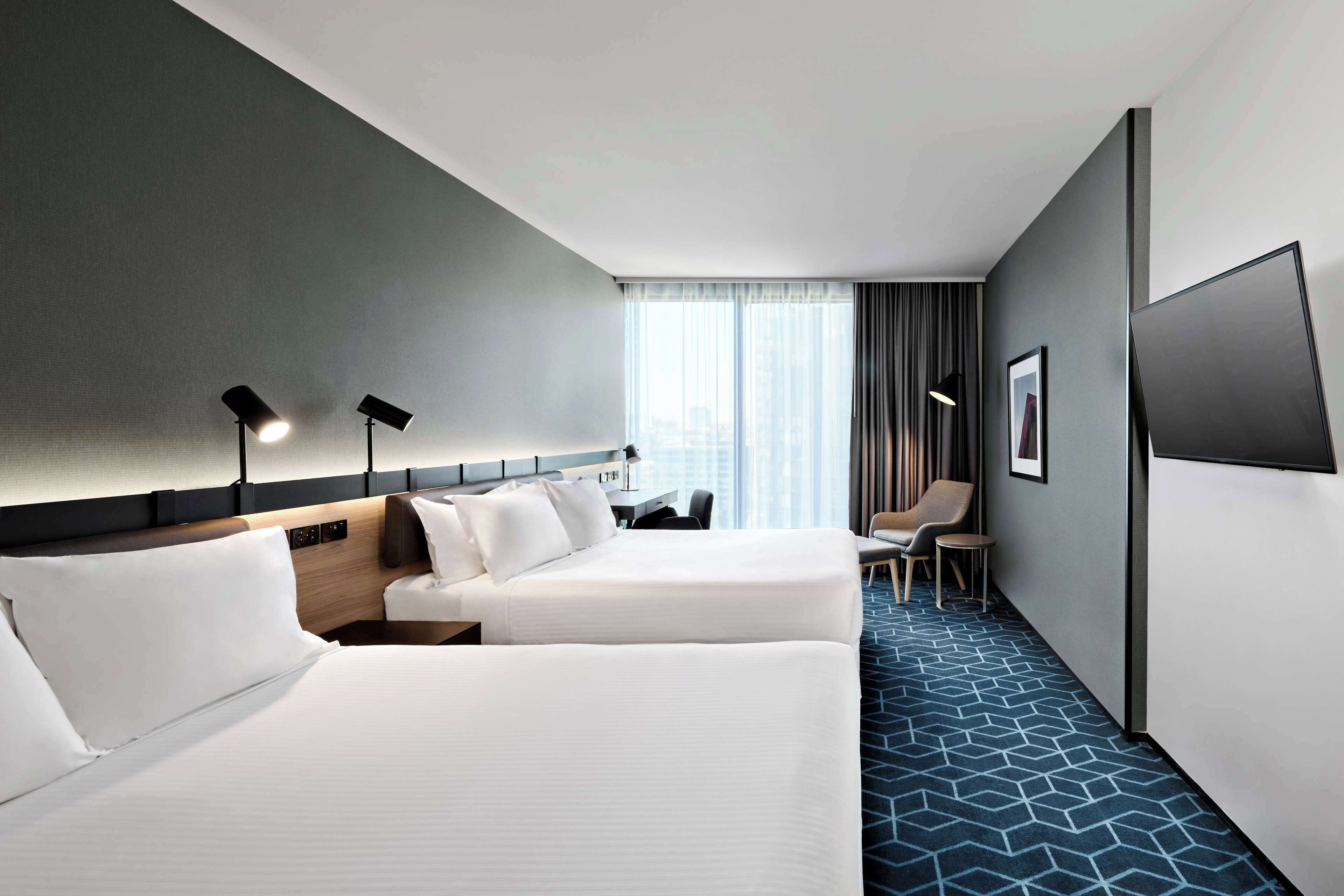 Habitación de hotel con dos camas, un escritorio, una silla, un televisor y un ventanal grande con cortinas.