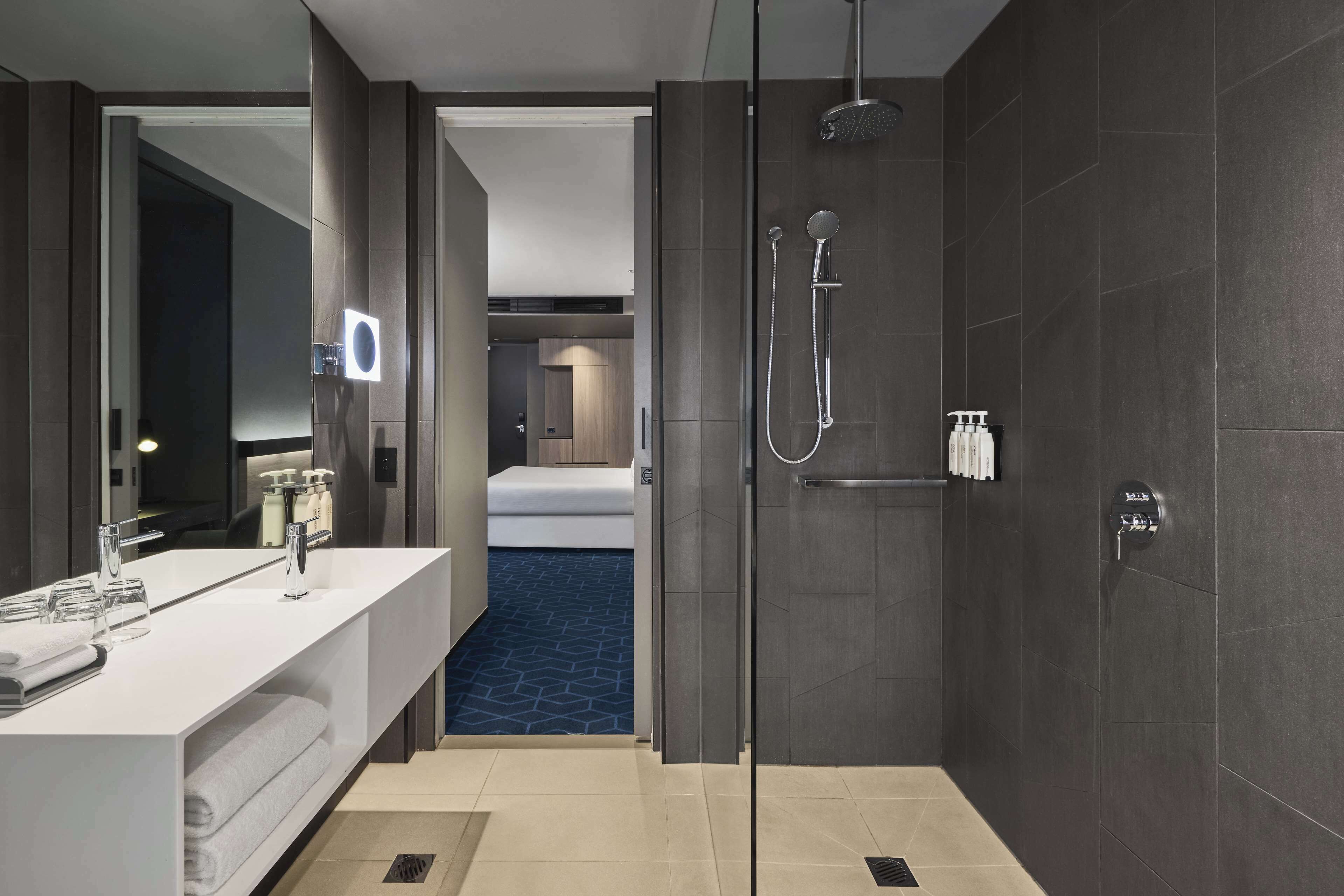 Un baño moderno con un espejo grande, zona de ducha y una habitación visible al fondo.
