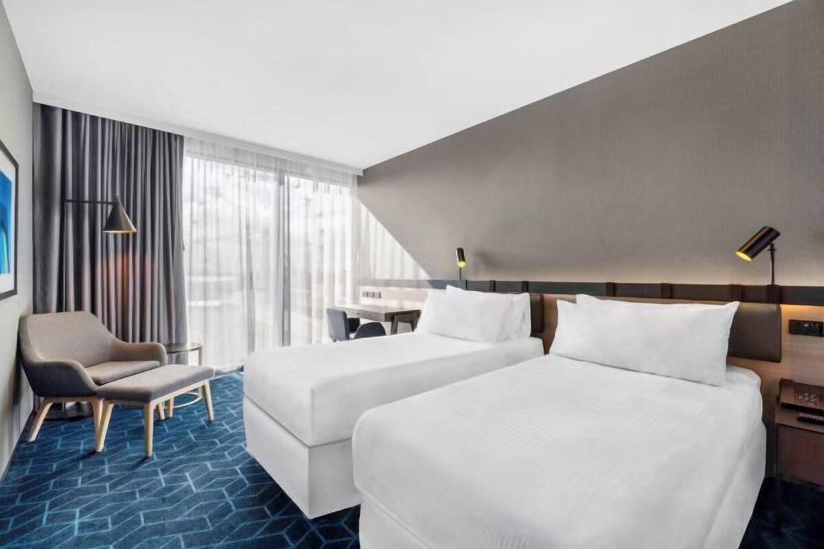 Ein Hotelzimmer mit zwei Betten, einem Schreibtisch, einem Stuhl und einem großen Fenster mit Vorhängen.
