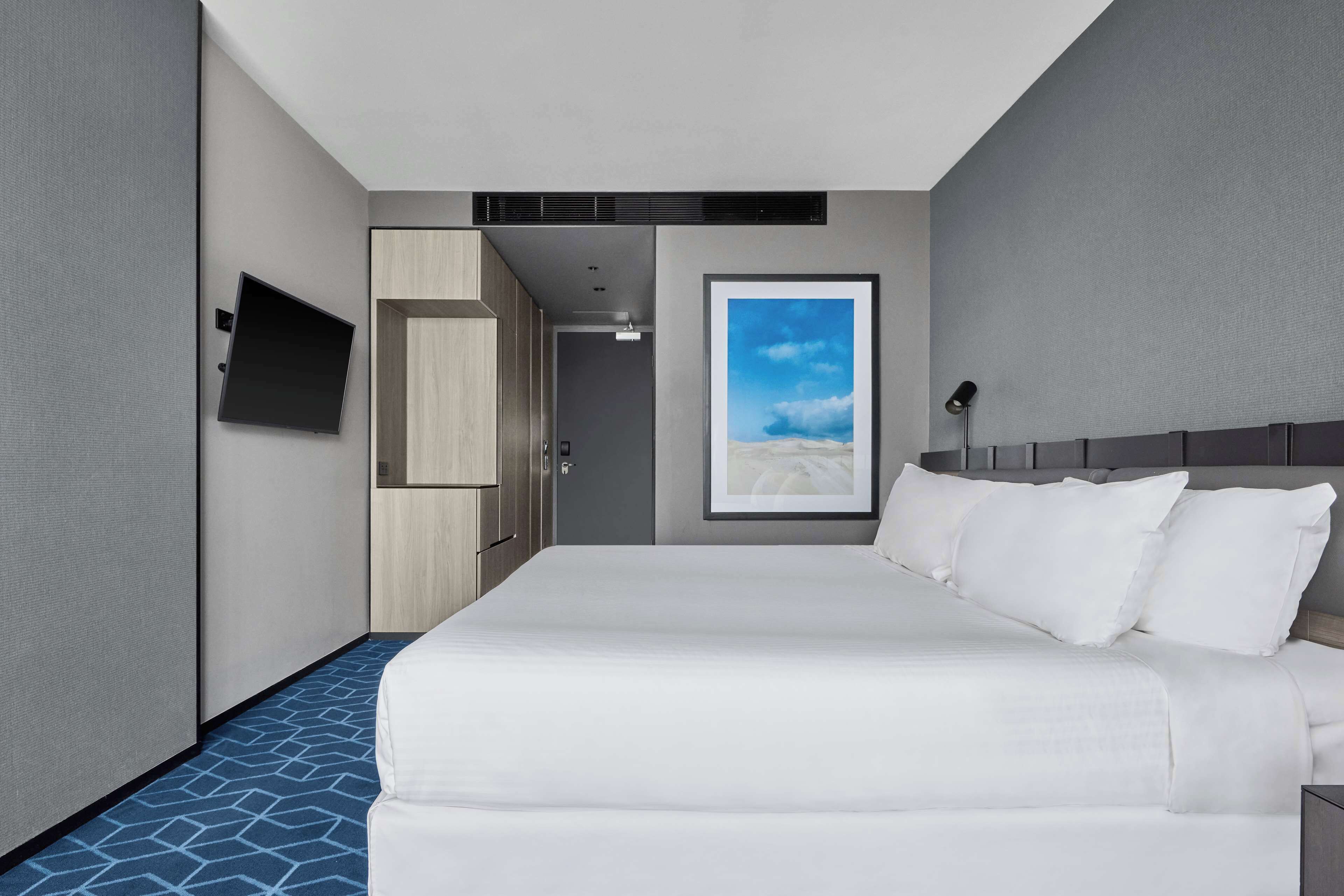 Una habitación de hotel con una cama grande, un televisor de pantalla plana montado en la pared, un cuadro enmarcado con una playa y una alfombra azul estampada.