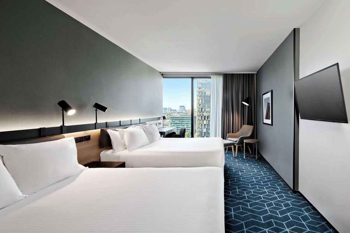 Ein Hotelzimmer mit zwei Betten, einem Schreibtisch, einem Sessel, einem Fernseher und einem großen Fenster mit Blick auf die Stadt.