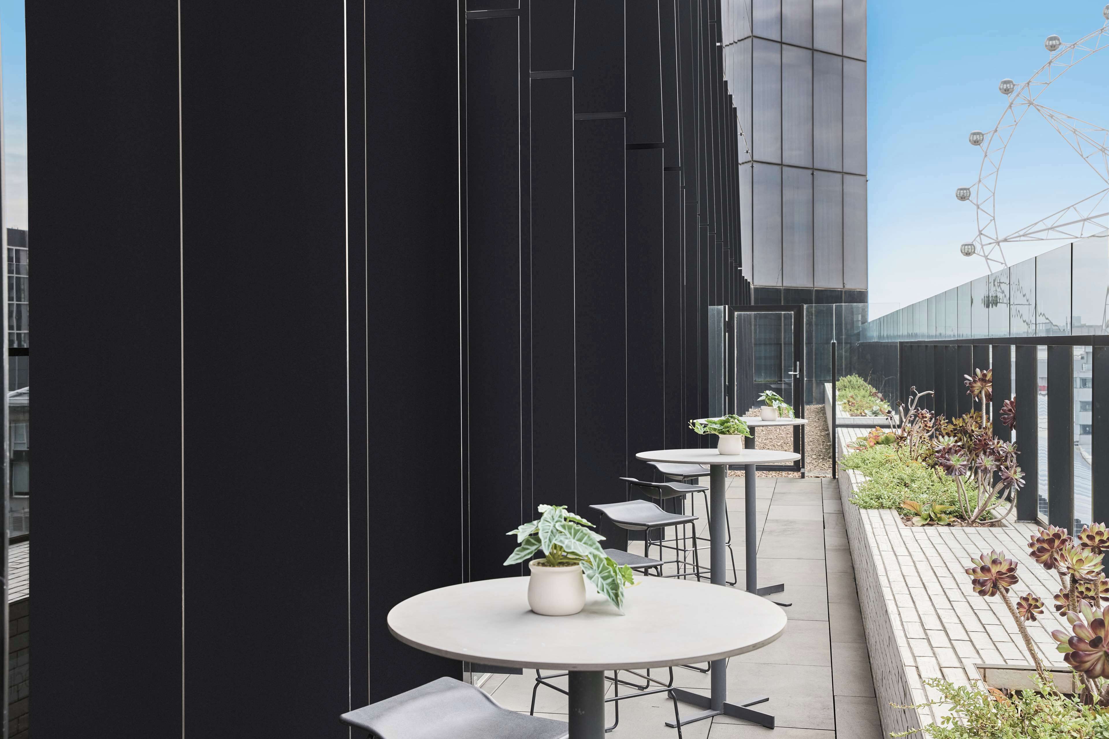 A rooftop terrace with round tables and chairs, potted plants, and a view of a Ferris wheel.