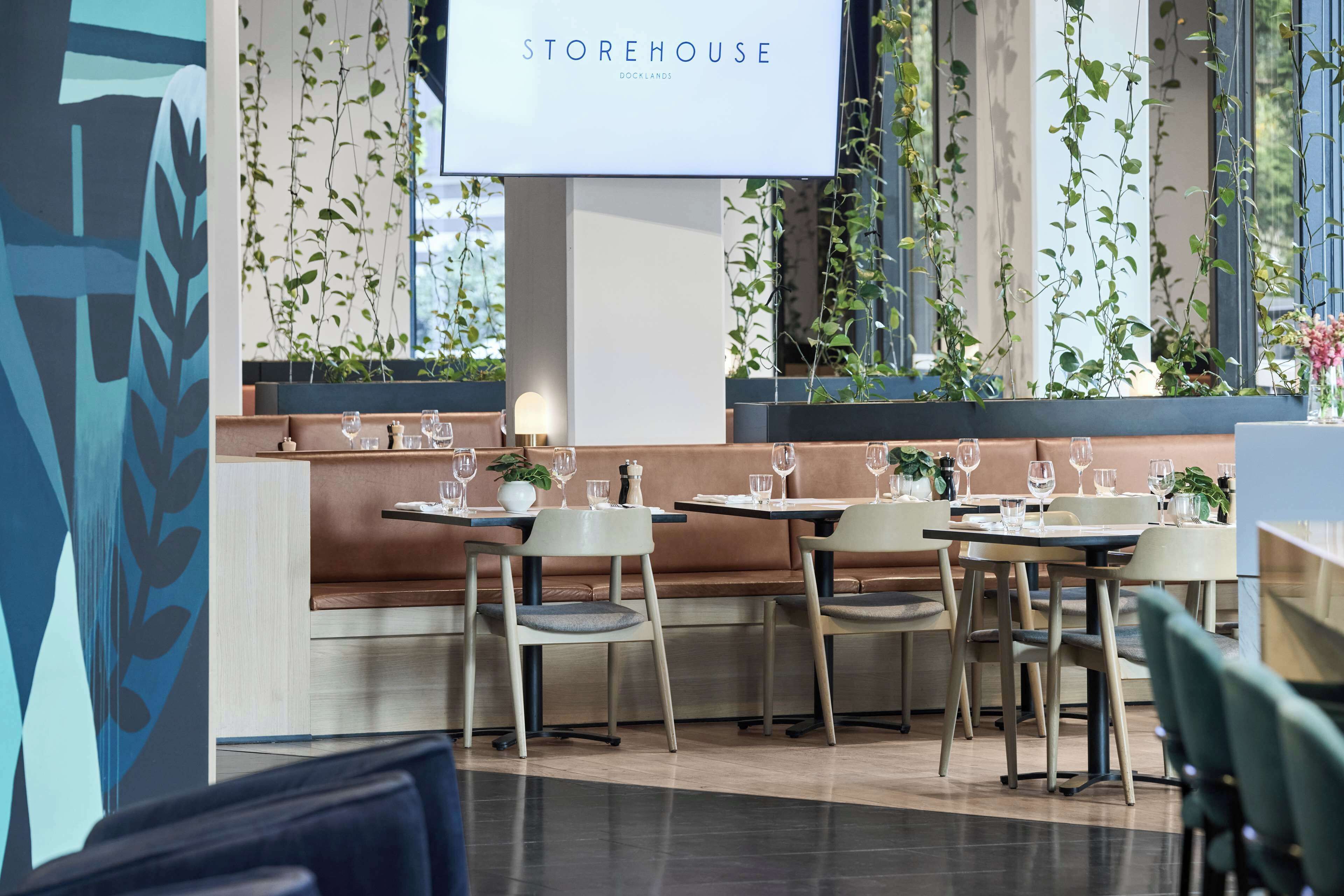 Un interior de restaurante moderno con un letrero 'STOREHOUSE', mesas de madera, asientos acolchados y plantas verdes.