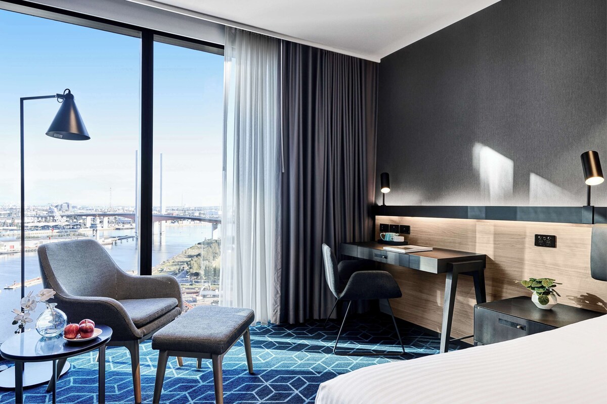 A hotel room with a large window, a desk, a chair, a bed, and a small table with a vase and fruit.
