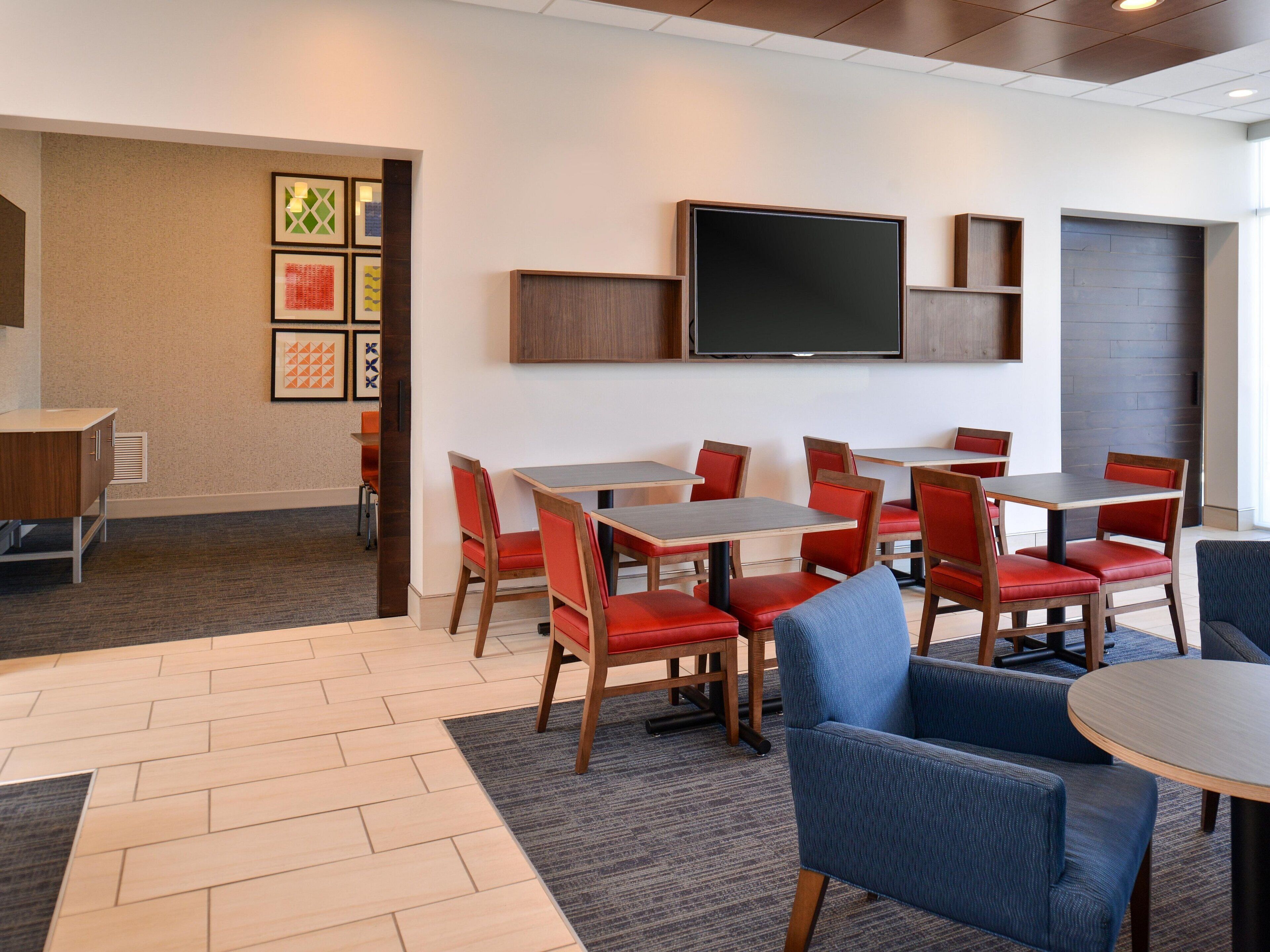 A hotel room with a dining area featuring tables and chairs, a flat-screen TV mounted on the wall, and a carpeted floor.