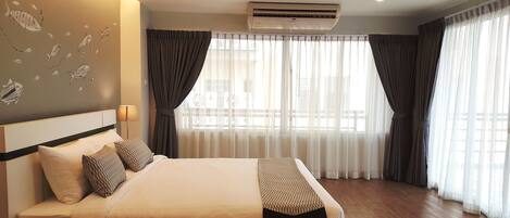 A hotel room with a large bed, a ceiling air conditioner, and sheer curtains on the windows.