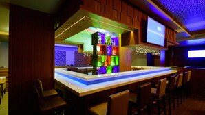 Una zona de bar con una barra alargada, varios taburetes y un estante iluminado y de colores.