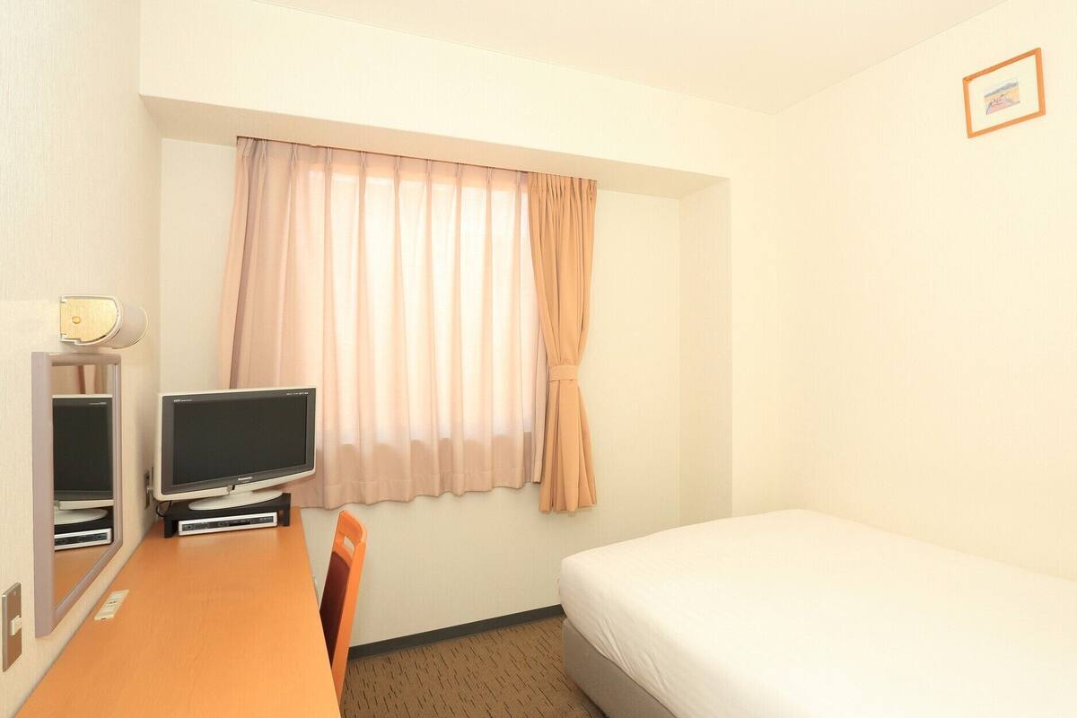 Ein Hotelzimmer mit einem Einzelbett, einem Schreibtisch mit Computer, einem Stuhl und einem Fenster mit Vorhängen.