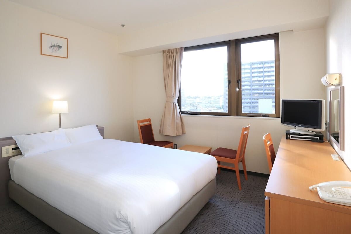 Ein Hotelzimmer mit einem Bett, einem Schreibtisch mit Computer und Telefon, einem Sessel, einem Fenster mit Vorhängen und einem Bild an der Wand.