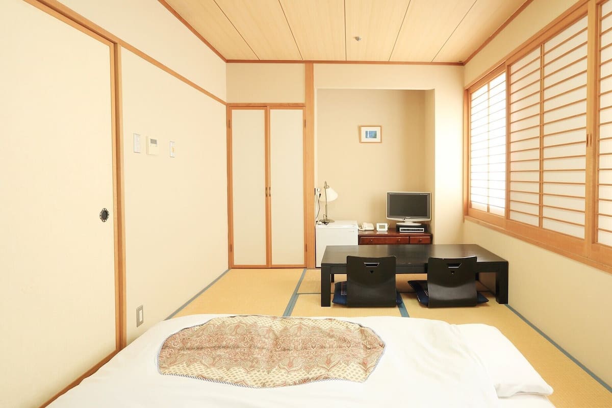 Ein traditioneller japanischer Raum mit Tatami-Bodenbelag, einem Bett, einem Schreibtisch mit Stuhl, einem Fernseher und Schiebetüren.