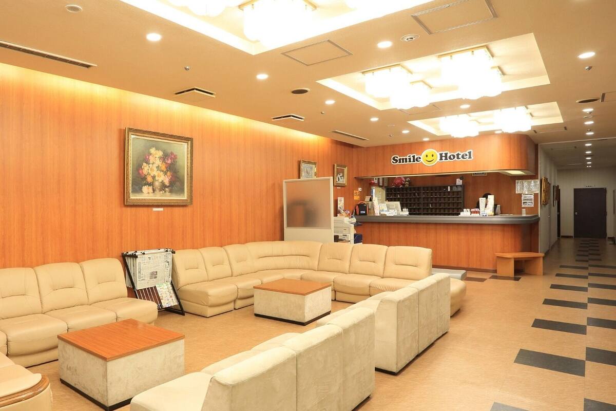 Ein Hotel-Empfangsbereich mit einer Holzwand, einem Empfangstresen, einer Sitzecke mit Sofas und Poufs sowie einem gerahmten Blumenbild.
