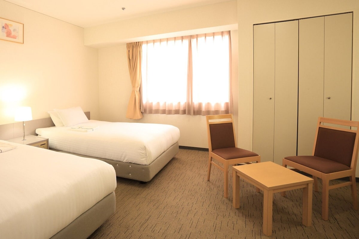 Ein Hotelzimmer mit zwei Betten, einem Schreibtisch, einem Stuhl, einem Kleiderschrank und einem Fenster mit Vorhängen.