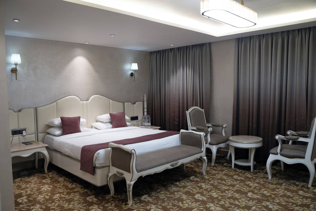 Une chambre d’hôtel avec un grand lit, deux tables de chevet, un bureau, une chaise, une table ronde et une grande fenêtre avec des rideaux.