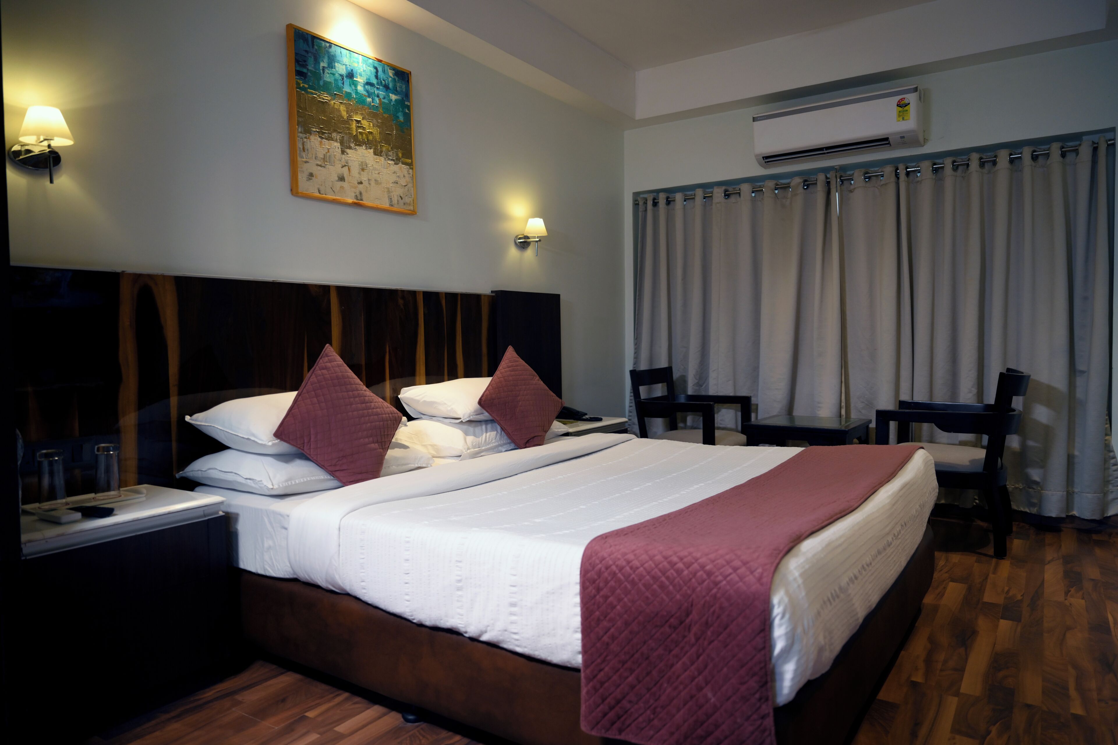 A hotel room with a large bed, two bedside tables, a desk, and a chair. There is a painting on the wall and an air conditioning unit above the bed.