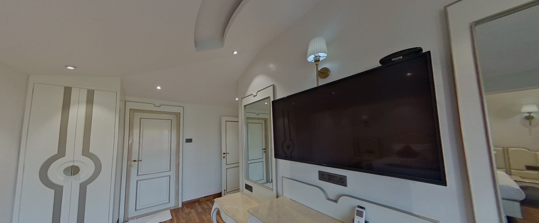 Una habitación de hotel moderna con un televisor de pantalla plana grande montado en la pared, un espejo y una lámpara en el techo.
