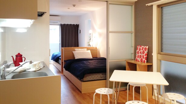 A compact hotel room with a bed, a small table, two stools, a sink with a red kettle, and a window with a view. - Stella Inn Karasuma Gojo (Kyoto)