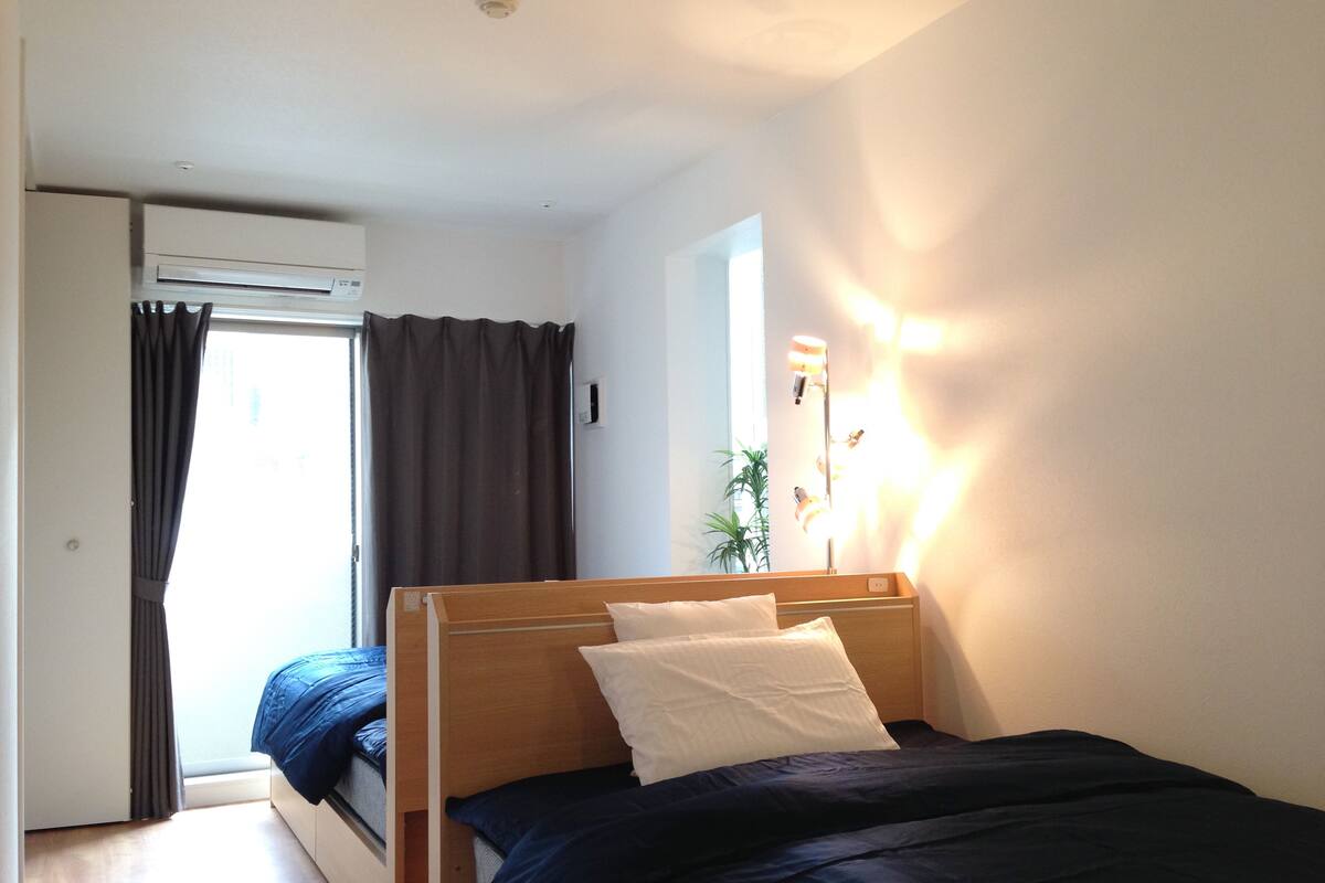 A bedroom with a bed, a window, a plant, and an air conditioning unit.