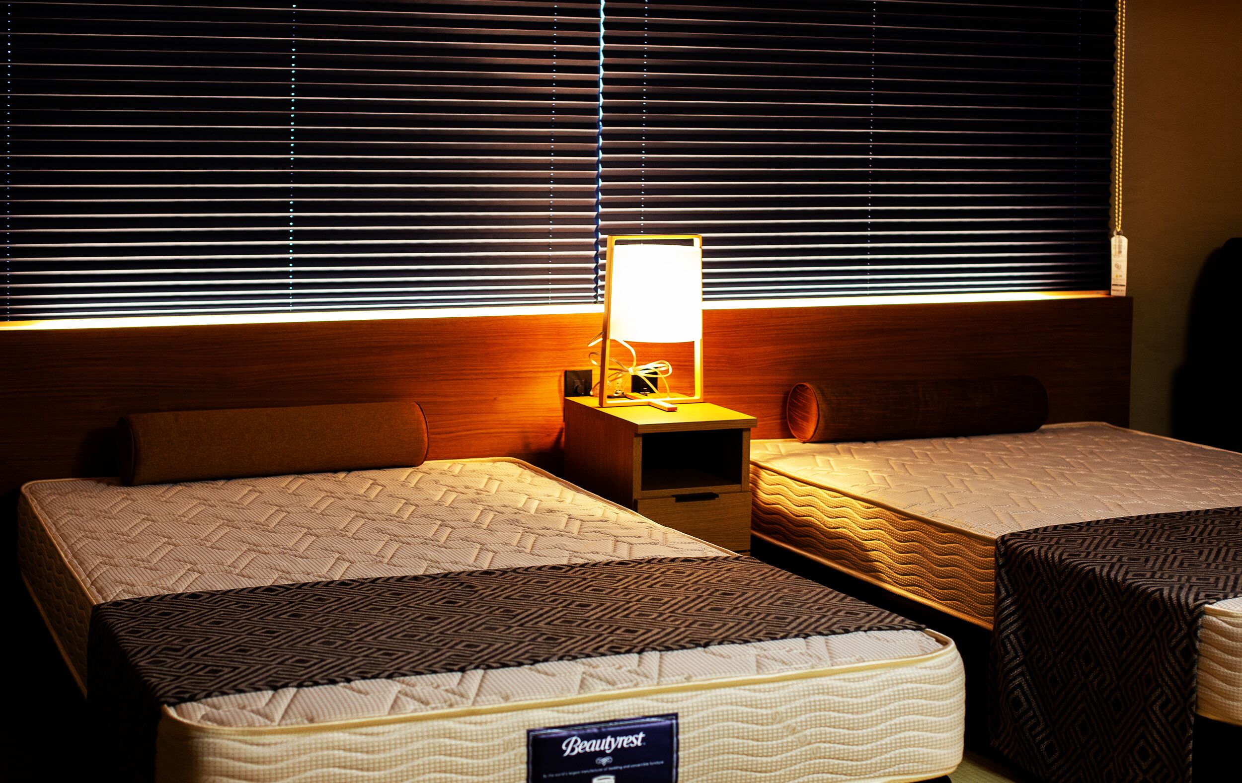 A hotel room with two beds, a nightstand with a lamp, and a window with blinds.
