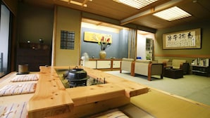Una habitación japonesa tradicional con suelo de tatami, una mesa de madera baja, cojines, un futón en el centro, un futón junto a la entrada, un armario de madera, una estantería con libros y una obra de caligrafía enmarcada en la pared.