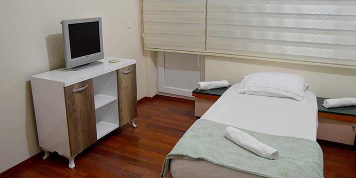 Une petite pièce simple, avec un lit, un bureau doté d’un ordinateur et une fenêtre avec des stores.