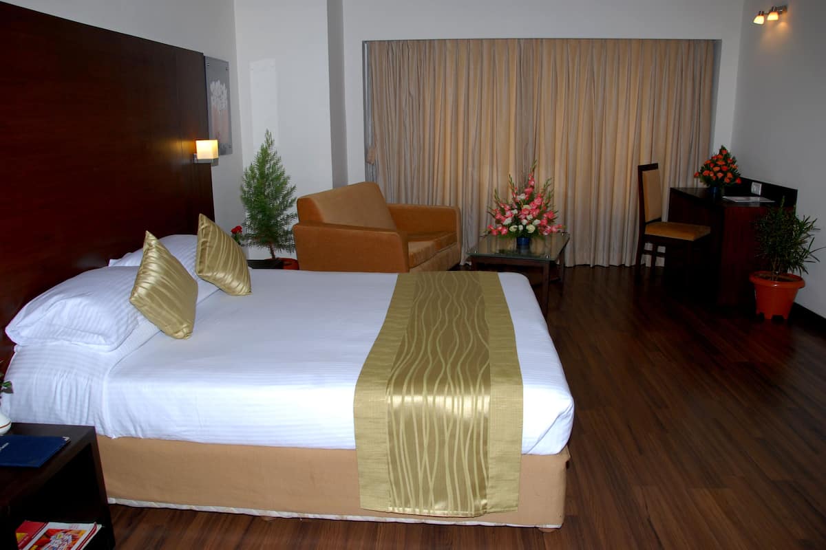 Habitación de hotel con una cama grande, cabecera de madera, una mesita con un jarrón de flores, una silla y un escritorio con una computadora.