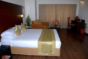A hotel room with a large bed, a wooden headboard, a small table with a vase of flowers, a chair, and a desk with a computer. - Hotel Siddharta (Mysore)