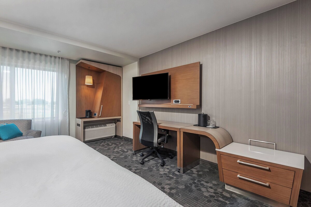 A hotel room with a bed, a desk with a chair, a television, and a small kitchenette area.