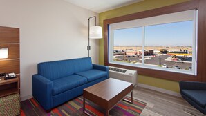 A hotel room with a blue sofa, a wooden coffee table, a lamp, and a large window with a view of a gas station and parking lot. - Holiday Inn Express & Suites Brigham City - North Utah by IHG (Brigham City)