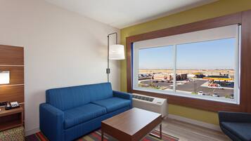 Habitación de hotel con sofá azul, una mesa de centro de madera, lámpara y ventana grande con vistas a una gasolinera y aparcamiento.