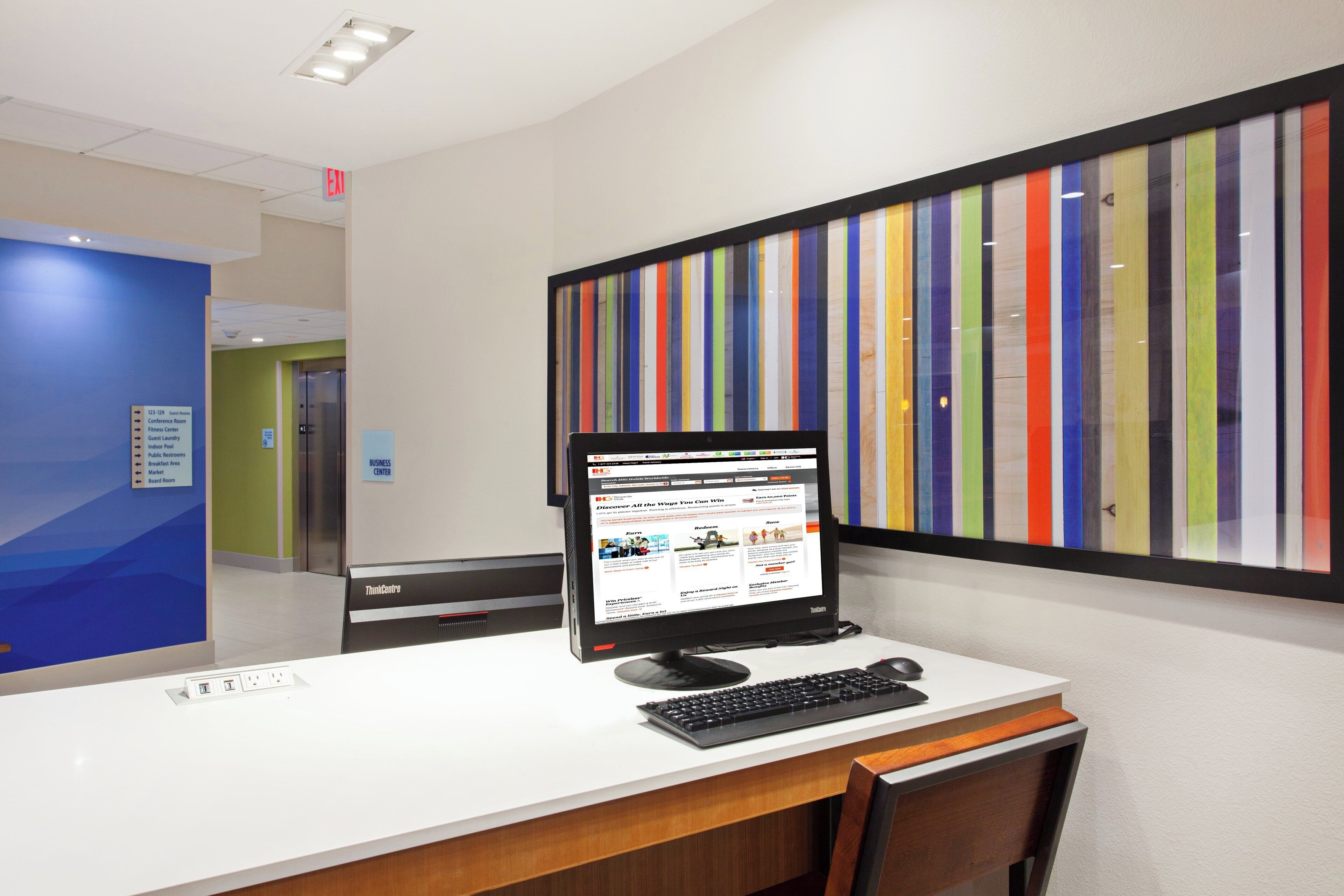 A hotel reception desk with a computer monitor displaying a website, a framed artwork with vertical stripes, and a sign indicating the business center.