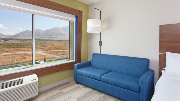 A room with a large window, a blue sofa, a bedside table, and a white lamp. - Holiday Inn Express & Suites Brigham City - North Utah by IHG (Brigham City)