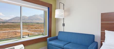 A room with a large window, a blue sofa, a bedside table, and a white lamp.
