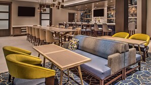 A modern hotel lobby with a long dining table, multiple chairs, a sofa, and a bar area. - Hyatt House Raleigh / RDU / Brier Creek (Raleigh)