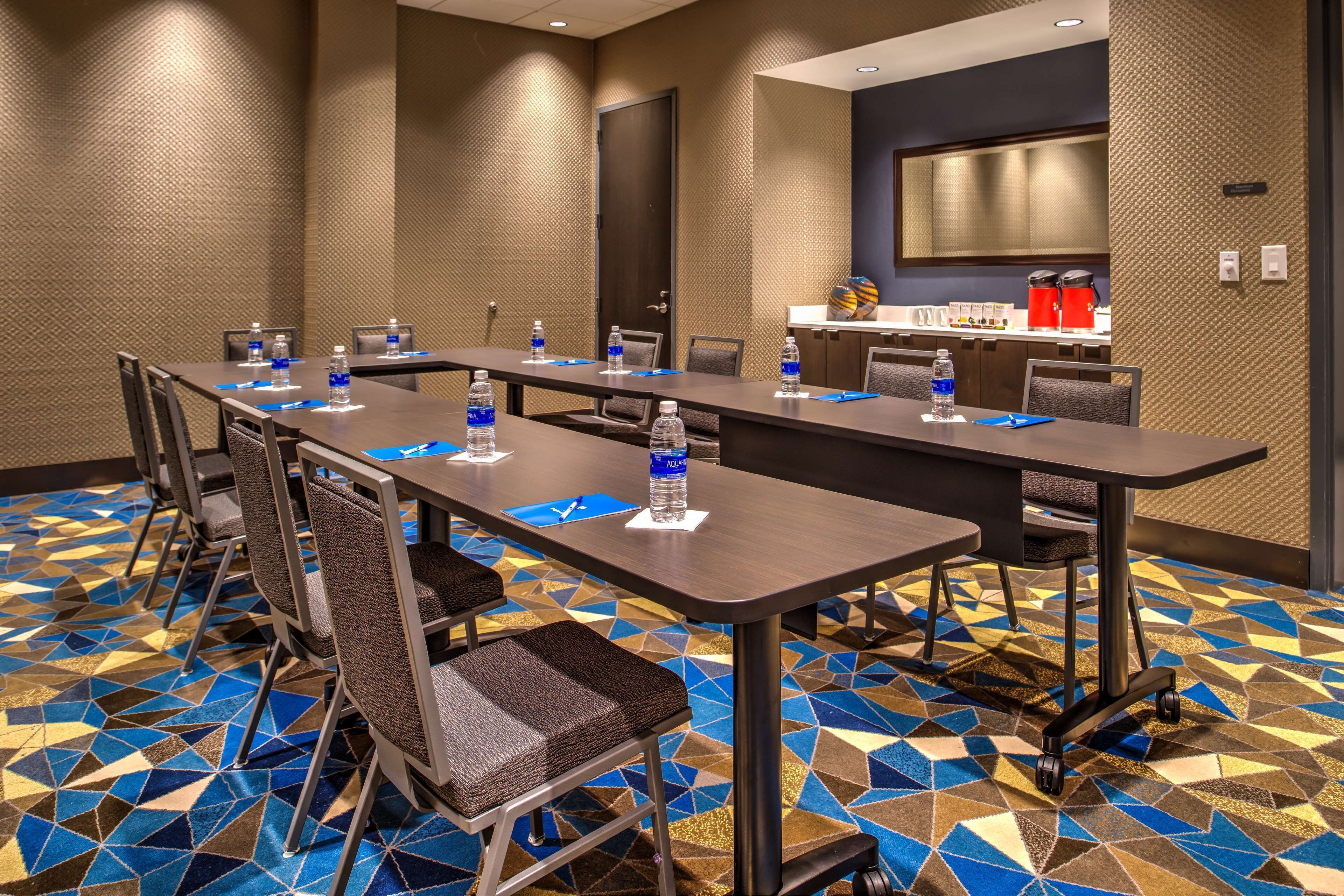 A conference room with a long table, chairs, water bottles, and a kitchen area in the background.