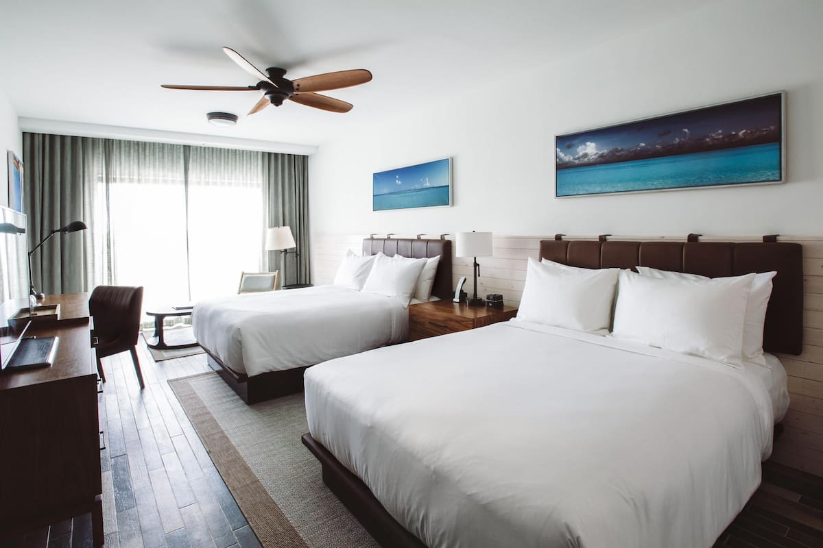 A hotel room with two beds, a desk, a chair, a ceiling fan, and two paintings on the wall.