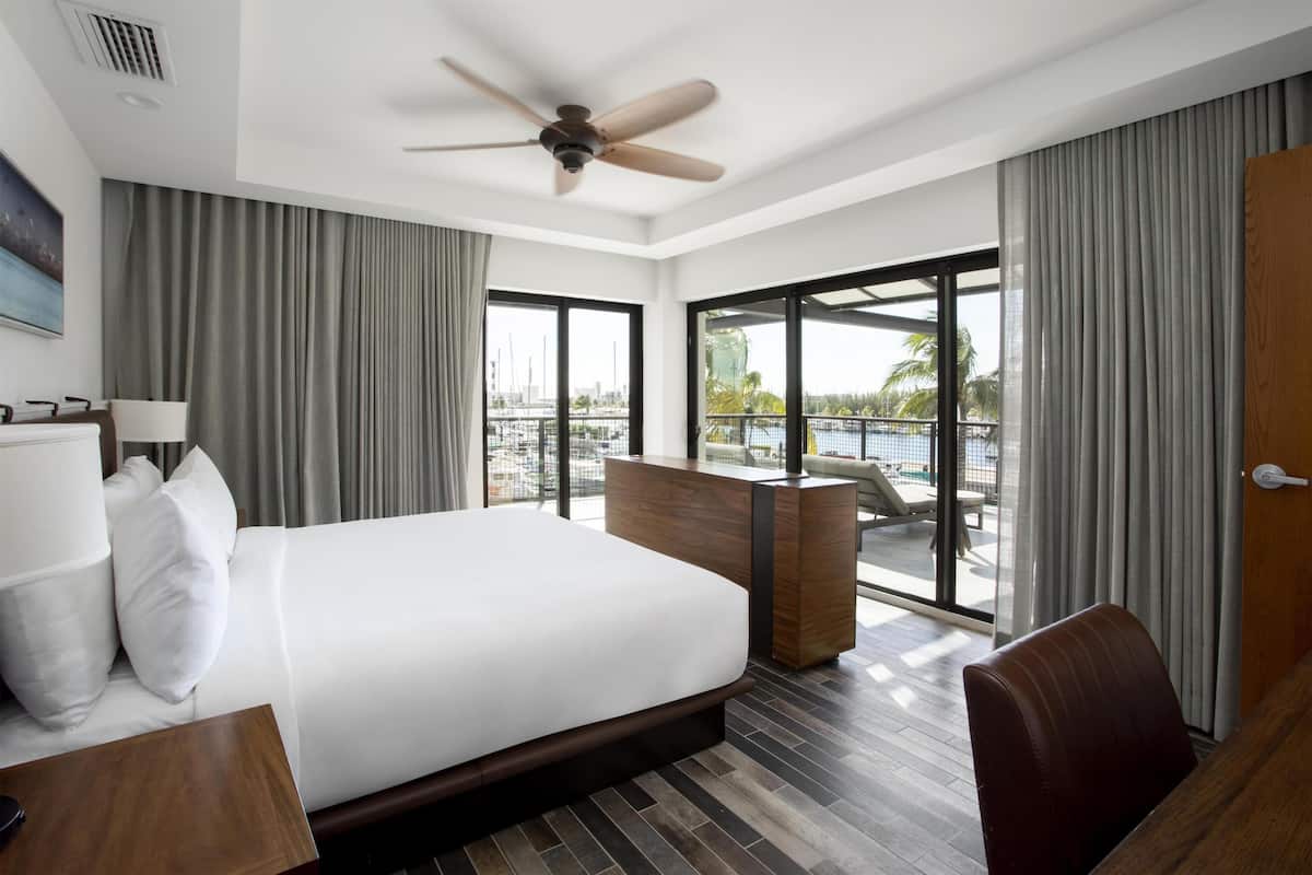 A modern hotel room with a large bed, a desk, a chair, a ceiling fan, and a view of a marina through large windows.