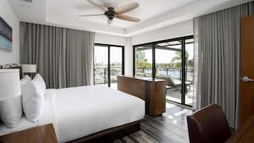 A modern hotel room with a large bed, a desk, a chair, a ceiling fan, and a view of a marina through large windows.